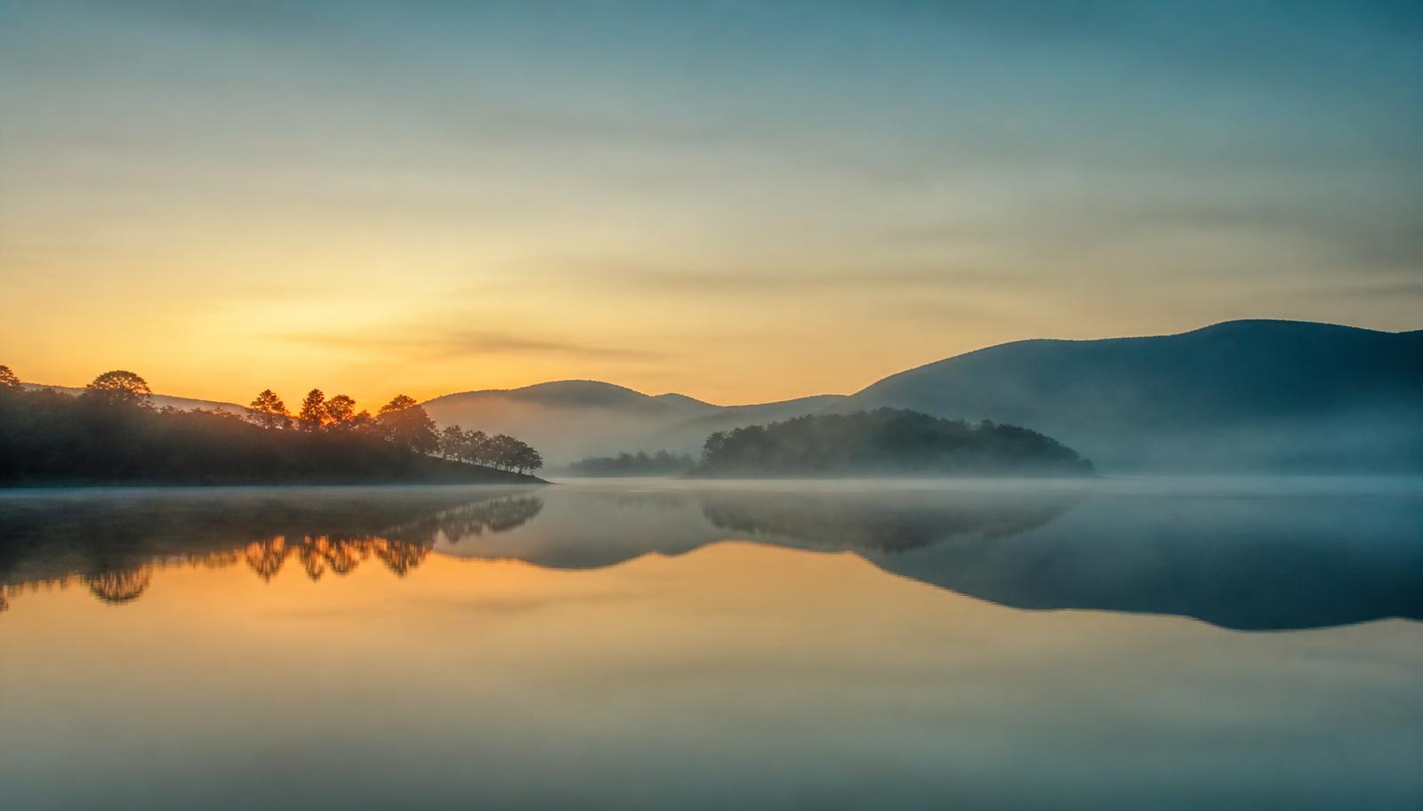 Lago tranquilo ao amanhecer, com névoa suave e céu dourado